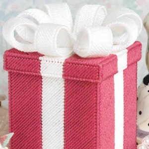 May include: A handmade pink and white gift box, topped with a large white bow. The box has a vertical white stripe down the front and is created using a cross-stitch method. The bow provides a decorative element.