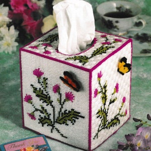 May include: White tissue box cover with a pink border and a floral design featuring green leaves and pink flowers. Two butterflies are attached to the cover, one orange and one yellow.