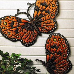 Puede incluir: Dos mariposas bordadas de color naranja y negro con detalles blancos montadas en un panel de madera blanco.