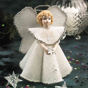 May include: A white beaded angel figurine with a halo and holding a star. The angel has a porcelain face with blonde hair and wears a white dress with silver trim. Decorative stars and a glass candle holder are in the background.
