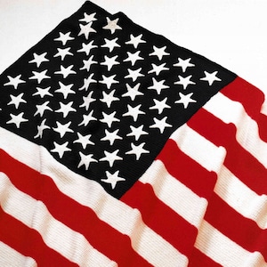 May include: A knitted American flag with a black field of stars and red and white stripes. The white stars are evenly spaced on the black field. The red and white stripes are alternating and run horizontally across the bottom of the flag.