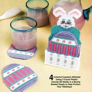 May include: A set of four colorful, stitched coasters in a bunny-shaped basket. The coasters feature a striped design in pink, purple, and white, with small flower accents. The text on the image reads: "4 Colorful Coasters Stitched Using 7-Count Plastic Canvas Sit Neatly in a Bunny Basket Ready to Help Protect Your Tabletops."