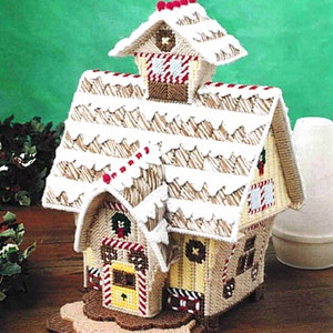 May include: A handcrafted Christmas village house with a snow-covered roof and decorative details. The house features a yellow door with red and white trim, and a small wreath. The roof is adorned with red accents.