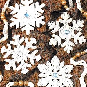 May include: Four white crocheted snowflake ornaments with blue gem centers. The snowflakes are arranged on a patterned brown and gold fabric, with white rope and gold bead accents. These handmade decorations are perfect for winter holiday decor.