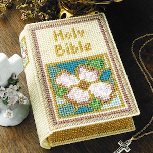 May include: A cream-colored, cross-stitched Holy Bible with gold lettering and a floral design. The cover features a pink and blue flower. A gold cross necklace rests nearby.