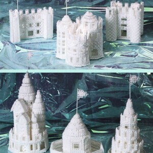 May include: White plastic canvas castle models with various architectural designs, including towers, turrets, and flags. The castles are displayed on a shiny, teal-colored surface. The text "produced by Jean Leinhauser" is visible.