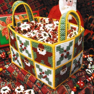 Puede incluir: Una cesta navideña hecha a mano llena de caramelos envueltos. La cesta está decorada con diseños de Papá Noel y acebo en rojo, verde y blanco. Las asas son verdes con ribetes amarillos. La cesta se encuentra sobre una tela roja estampada festiva.