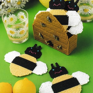 May include: Set of four yellow and black bee-shaped coasters with white wings. The coasters are arranged in a beehive-shaped holder. The holder is made of brown yarn and has a small opening at the bottom. The coasters are on a green surface.
