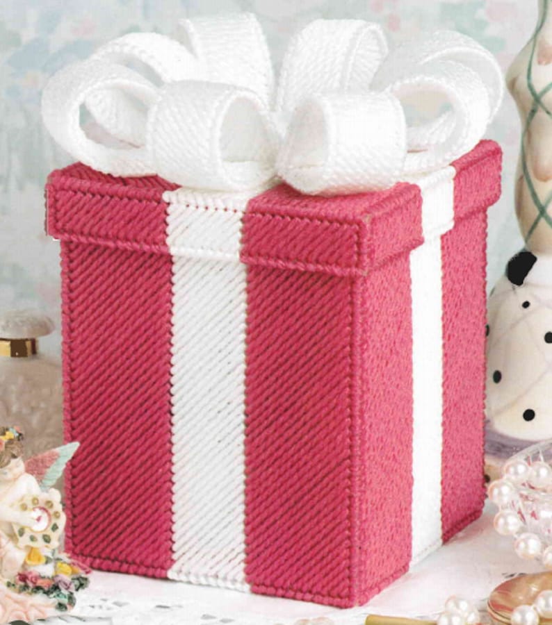 May include: A handcrafted, pink and white gift box with a large white bow on top. The box features a vertical white stripe down the front and is made with a cross-stitch technique. The bow adds a decorative touch.
