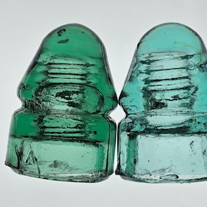 May include: Two antique glass insulators, one emerald green and the other teal, are displayed side-by-side. The insulators have a distinctive shape with a rounded top and a grooved body. The green insulator has an "A" etched on its base. These vintage pieces are likely from a telegraph or power line.