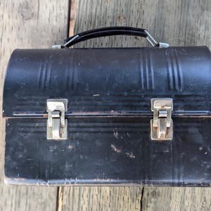 May include: A vintage black lunchbox with a curved top and vertical embossed lines. The lunchbox has a black handle and two silver latches. The lunchbox is sitting on a weathered wooden surface.