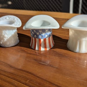 May include: Three white miniature top hats. One hat has a textured, patterned design, another features a patriotic red, white, and blue design, and the third has a smooth, ribbed texture. The hats are displayed on a wooden surface.