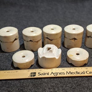 May include: A collection of off-white ceramic insulators, some with cracks, arranged on a gray surface. The insulators are cylindrical with a hole in the center. A wooden ruler with text is in the foreground.
