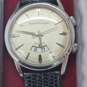 K&ouml;nnte beinhalten: Eine Vintage-Armbanduhr von Girard-Perregaux mit silberfarbenem Geh&auml;use und cremefarbenem Zifferblatt. Die Uhr hat silberne Zeiger und Markierungen, eine Datumsanzeige und ein schwarzes Lederarmband. Die Uhr hat zwei Kronen und der Text "Girard-Perregaux" ist sichtbar.