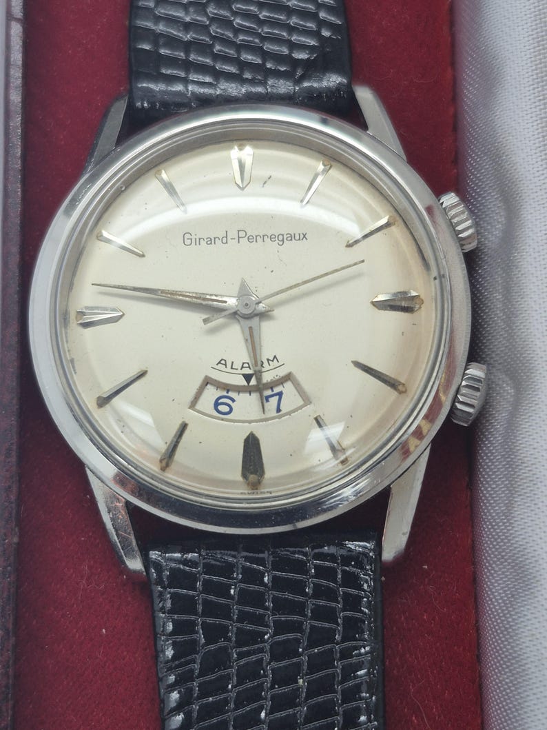 K&ouml;nnte beinhalten: Eine Vintage-Armbanduhr von Girard-Perregaux mit silberfarbenem Geh&auml;use und cremefarbenem Zifferblatt. Die Uhr hat silberne Zeiger und Markierungen, eine Datumsanzeige und ein schwarzes Lederarmband. Die Uhr hat zwei Kronen und der Text "Girard-Perregaux" ist sichtbar.