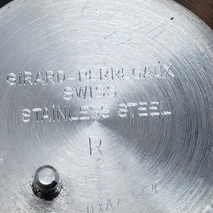 K&ouml;nnte beinhalten: Nahaufnahme eines Edelstahl-Uhrenr&uuml;ckens. Der Uhrenr&uuml;cken tr&auml;gt die Gravur "GIRARD-PERREGAUX SWISS STAINLESS STEEL R". Es gibt auch andere Markierungen und ein kleines Metallst&uuml;ck.