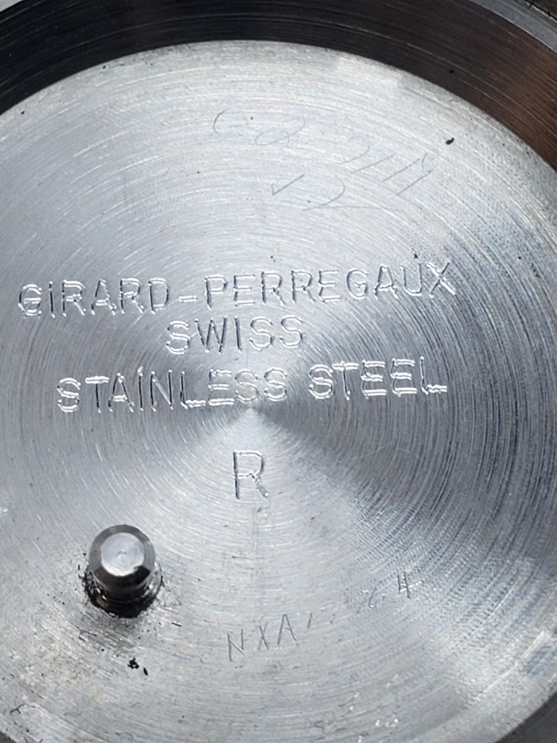 K&ouml;nnte beinhalten: Nahaufnahme eines Edelstahl-Uhrenr&uuml;ckens. Der Uhrenr&uuml;cken tr&auml;gt die Gravur "GIRARD-PERREGAUX SWISS STAINLESS STEEL R". Es gibt auch andere Markierungen und ein kleines Metallst&uuml;ck.