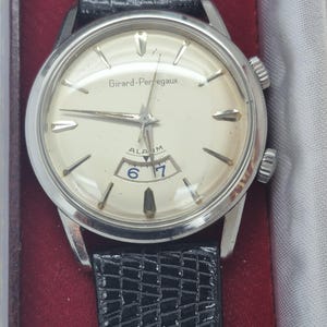 K&ouml;nnte beinhalten: Eine Vintage Girard-Perregaux Alarmuhr mit silberfarbenem Geh&auml;use und cremefarbenem Zifferblatt. Die Uhr hat schwarze Zeiger und Markierungen, ein Datumsfenster und ein schwarzes Lederarmband. Auf dem Zifferblatt steht "Girard-Perregaux" und "ALARM".