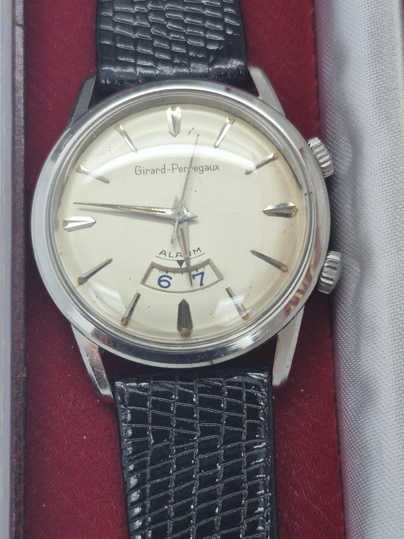K&ouml;nnte beinhalten: Eine Vintage Girard-Perregaux Alarmuhr mit silberfarbenem Geh&auml;use und cremefarbenem Zifferblatt. Die Uhr hat schwarze Zeiger und Markierungen, ein Datumsfenster und ein schwarzes Lederarmband. Auf dem Zifferblatt steht "Girard-Perregaux" und "ALARM".