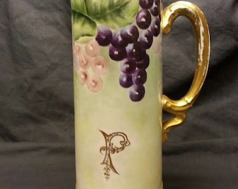 May include: Tall, cylindrical pitcher with a gold handle and base trim. The pitcher is painted with purple and pink grapes and green leaves. A gold-colored monogram is on the front.