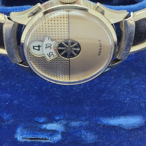 May include: A gold-toned Benrus watch with a black leather band. The watch face is divided into two sections, one with a rotating starburst design and the other with a date display showing the number 4. The watch is set against a blue velvet background.