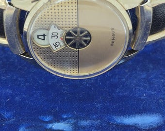Puede incluir: Un reloj Benrus dorado con una correa de cuero negro. La esfera del reloj está dividida en dos secciones, una con un diseño de estrella giratoria y otra con una pantalla de fecha que muestra el número 4. El reloj se presenta sobre un fondo de terciopelo azul.
