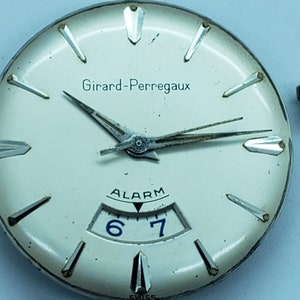 K&ouml;nnte beinhalten: Nahaufnahme eines Vintage-Zifferblatts von Girard-Perregaux. Die Uhr hat ein cremefarbenes Zifferblatt mit silberfarbenen Stundenmarkierungen und Zeigern. Das Wort "Girard-Perregaux" ist oben aufgedruckt. Die Uhr verf&uuml;gt au&szlig;erdem &uuml;ber eine Alarmfunktion mit den Zahlen 6 und 7.