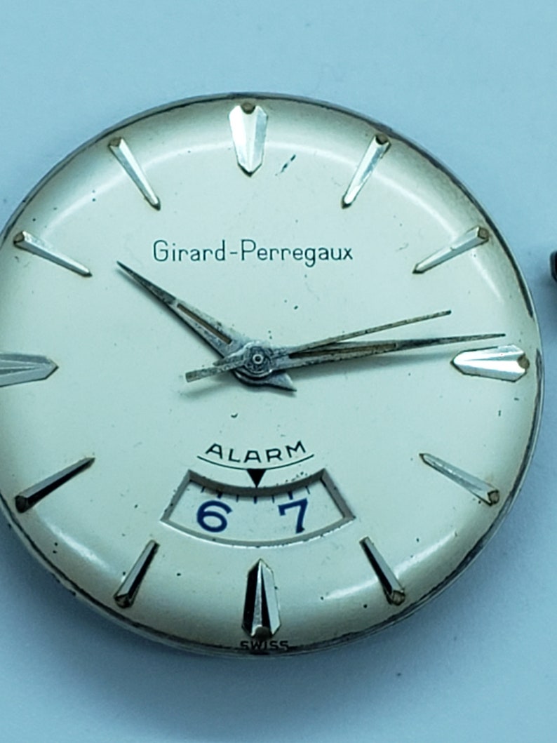 K&ouml;nnte beinhalten: Nahaufnahme eines Vintage-Zifferblatts von Girard-Perregaux. Die Uhr hat ein cremefarbenes Zifferblatt mit silberfarbenen Stundenmarkierungen und Zeigern. Das Wort "Girard-Perregaux" ist oben aufgedruckt. Die Uhr verf&uuml;gt au&szlig;erdem &uuml;ber eine Alarmfunktion mit den Zahlen 6 und 7.