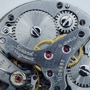 K&ouml;nnte beinhalten: Nahaufnahme eines Vintage-Uhrwerks, das komplizierte Zahnr&auml;der, Schrauben und Juwelen zeigt. Die Metallkomponenten sind silberfarben, mit goldfarbenen Zahnr&auml;dern und roten Juwelen. Das Uhrwerk ist mit "Girard-Perregaux", "17 Jewels" und "Swiss" beschriftet.