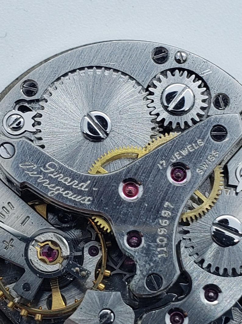 K&ouml;nnte beinhalten: Nahaufnahme eines Vintage-Uhrwerks, das komplizierte Zahnr&auml;der, Schrauben und Juwelen zeigt. Die Metallkomponenten sind silberfarben, mit goldfarbenen Zahnr&auml;dern und roten Juwelen. Das Uhrwerk ist mit "Girard-Perregaux", "17 Jewels" und "Swiss" beschriftet.