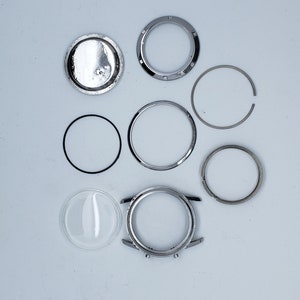 K&ouml;nnte beinhalten: Eine Ansammlung von Uhrenkomponenten, darunter ein silbernes Uhrengeh&auml;use, ein klares Glas, verschiedene silberne Ringe und eine R&uuml;ckseite. Die Teile sind auf einer hellgrauen Oberfl&auml;che angeordnet, was auf eine Uhrenreparatur oder -restaurierung hindeutet.