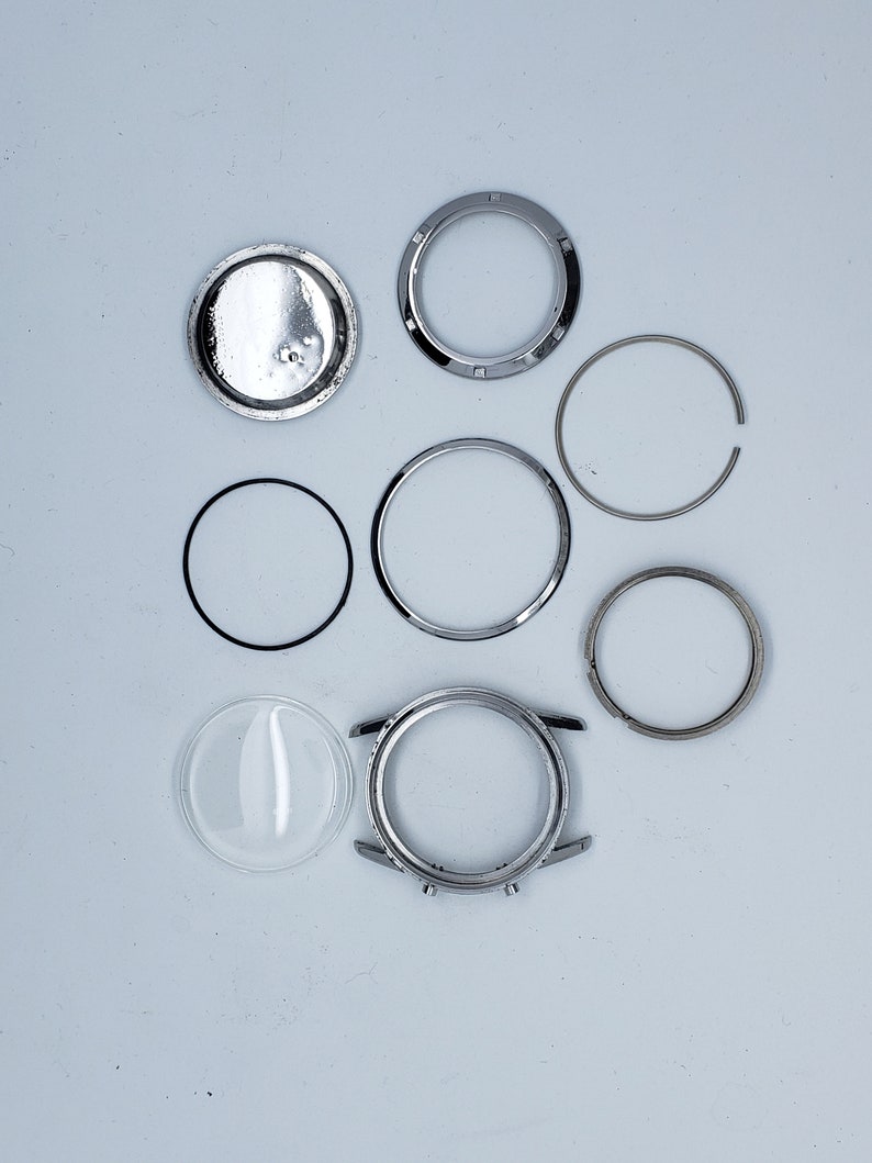 K&ouml;nnte beinhalten: Eine Ansammlung von Uhrenkomponenten, darunter ein silbernes Uhrengeh&auml;use, ein klares Glas, verschiedene silberne Ringe und eine R&uuml;ckseite. Die Teile sind auf einer hellgrauen Oberfl&auml;che angeordnet, was auf eine Uhrenreparatur oder -restaurierung hindeutet.