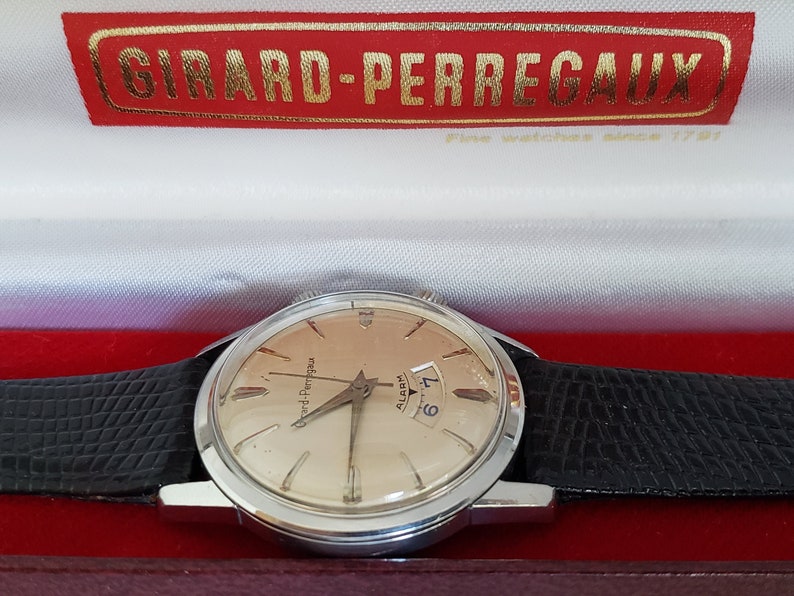 K&ouml;nnte beinhalten: Eine Vintage-Girard-Perregaux-Uhr mit silbernem Geh&auml;use und hellem Zifferblatt, mit Datumsanzeige. Die Uhr hat ein schwarzes Lederarmband und wird in einer rot-wei&szlig;en Schachtel mit dem Markennamen in goldfarbenen Buchstaben pr&auml;sentiert.