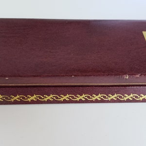 K&ouml;nnte beinhalten: Eine weinrote Schachtel mit einem goldenen Zierrand und einem goldenen Logo in der oberen rechten Ecke. Die Schachtel hat eine leicht strukturierte Oberfl&auml;che und scheint aus Leder oder einem &auml;hnlichen Material zu bestehen. Die Schachtel ist geschlossen.