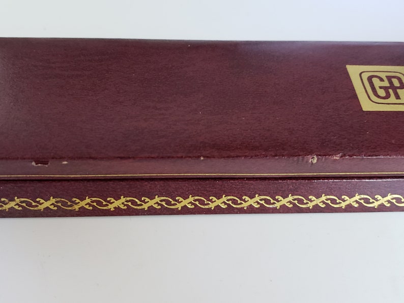 K&ouml;nnte beinhalten: Eine weinrote Schachtel mit einem goldenen Zierrand und einem goldenen Logo in der oberen rechten Ecke. Die Schachtel hat eine leicht strukturierte Oberfl&auml;che und scheint aus Leder oder einem &auml;hnlichen Material zu bestehen. Die Schachtel ist geschlossen.