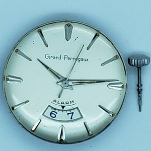 K&ouml;nnte beinhalten: Nahaufnahme eines Vintage-Zifferblatts der Girard-Perregaux-Uhr mit Zeigern, die die Uhrzeit anzeigen. Das cremefarbene Zifferblatt hat silberne Akzente und das Wort "ALARM" &uuml;ber den Zahlen 6 und 7. Zwei Uhrenkronen befinden sich auf beiden Seiten.