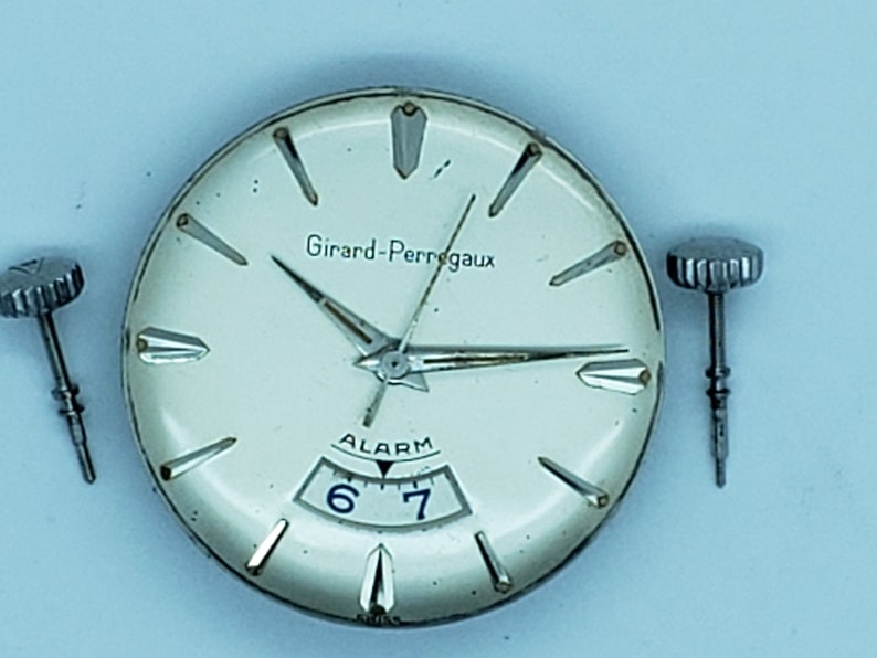 K&ouml;nnte beinhalten: Nahaufnahme eines Vintage-Zifferblatts der Girard-Perregaux-Uhr mit Zeigern, die die Uhrzeit anzeigen. Das cremefarbene Zifferblatt hat silberne Akzente und das Wort "ALARM" &uuml;ber den Zahlen 6 und 7. Zwei Uhrenkronen befinden sich auf beiden Seiten.
