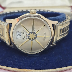May include: A gold-toned Benrus wristwatch with a compass-style design on the face. The watch features a date window and a gold-colored band. The watch is displayed in a blue velvet lined box.