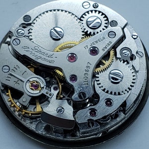 K&ouml;nnte beinhalten: Nahaufnahme eines Uhrwerks mit sichtbaren Zahnr&auml;dern, Juwelen und der Aufschrift "Girard-Perregaux". Der Uhrenmechanismus weist komplizierte Details auf, darunter kleine Schrauben und eine Seriennummer. Die W&ouml;rter "17 Jewels" und "Swiss" sind ebenfalls sichtbar.