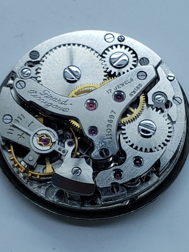 K&ouml;nnte beinhalten: Nahaufnahme eines Uhrwerks mit sichtbaren Zahnr&auml;dern, Juwelen und der Aufschrift "Girard-Perregaux". Der Uhrenmechanismus weist komplizierte Details auf, darunter kleine Schrauben und eine Seriennummer. Die W&ouml;rter "17 Jewels" und "Swiss" sind ebenfalls sichtbar.
