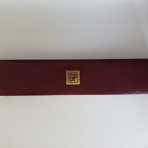 K&ouml;nnte beinhalten: Eine rechteckige, weinrote Schachtel mit einem goldfarbenen quadratischen Emblem in der Mitte. Das Emblem zeigt die Buchstaben "GP". Die Schachtel hat eine strukturierte Oberfl&auml;che und scheint aus Leder oder einem &auml;hnlichen Material zu bestehen.