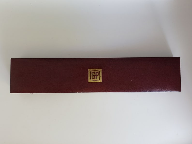K&ouml;nnte beinhalten: Eine rechteckige, weinrote Schachtel mit einem goldfarbenen quadratischen Emblem in der Mitte. Das Emblem zeigt die Buchstaben "GP". Die Schachtel hat eine strukturierte Oberfl&auml;che und scheint aus Leder oder einem &auml;hnlichen Material zu bestehen.