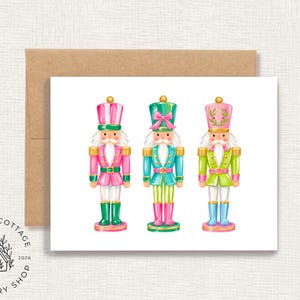 May include: A greeting card featuring a watercolor illustration of three nutcracker figures in pink, teal, and lime green uniforms with gold accents, positioned above a brown kraft paper envelope on a textured white background.