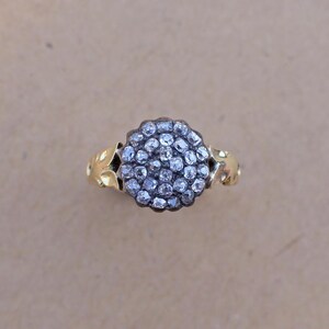 May include: A gold ring featuring a cluster of small, clear gemstones in a circular arrangement. The ring's band has a decorative, leaf-like design on the sides. The gemstones are set in a dark metal, creating a contrast with the gold band.