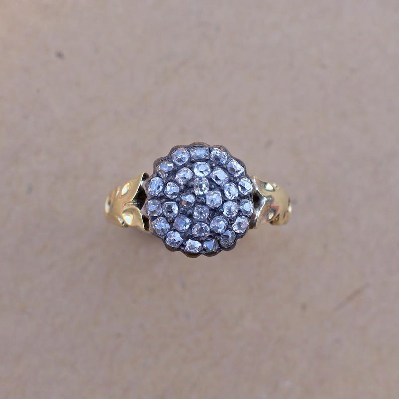 May include: A gold ring featuring a cluster of small, clear gemstones in a circular arrangement. The ring's band has a decorative, leaf-like design on the sides. The gemstones are set in a dark metal, creating a contrast with the gold band.