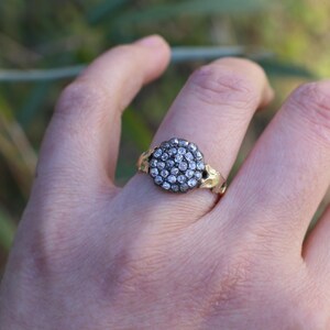 May include: A gold-toned ring with a cluster of small, clear gemstones set in a circular design. The ring's band features a decorative, leaf-like pattern on the sides, adding a touch of nature-inspired elegance to the jewelry.