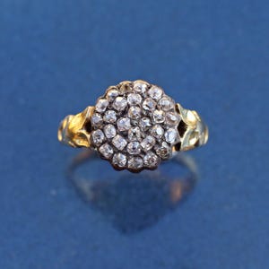 May include: A gold ring with a cluster of small, clear gemstones. The gemstones are set closely together, forming a rounded shape. The ring's band has a textured design, and the background is a solid blue.