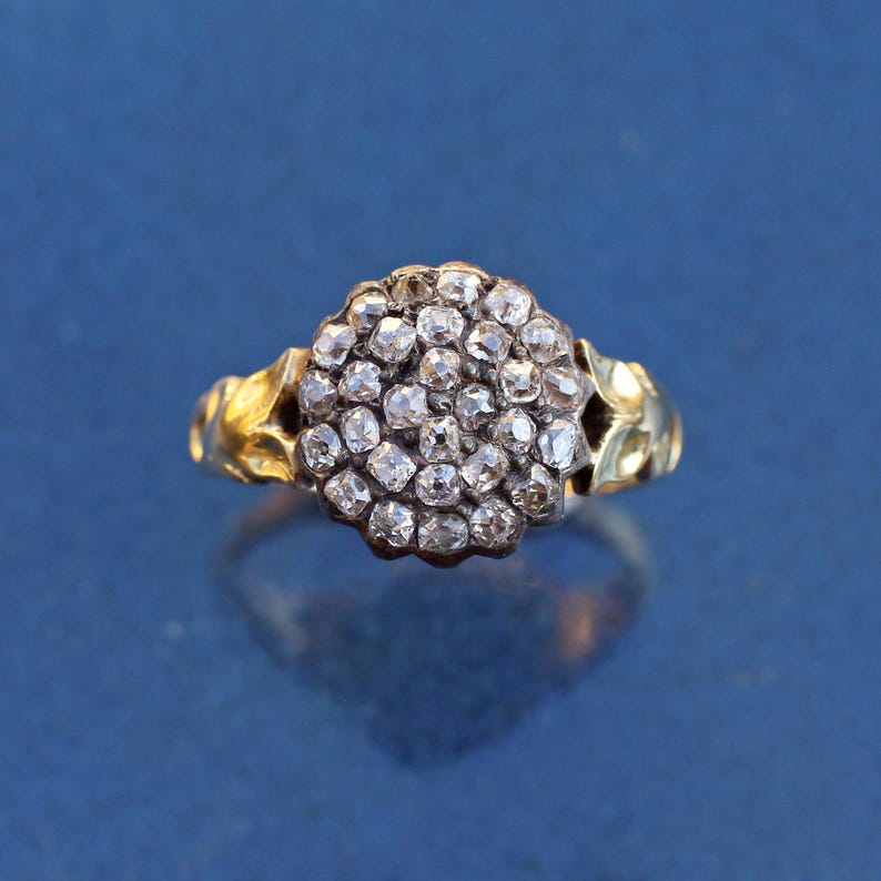 May include: A gold ring with a cluster of small, clear gemstones. The gemstones are set closely together, forming a rounded shape. The ring's band has a textured design, and the background is a solid blue.