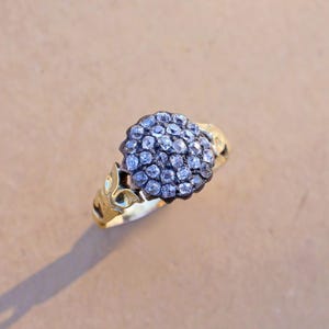 May include: A gold ring featuring a cluster of small, round, clear gemstones. The ring's band has a decorative floral design. The gemstones are set in a dark metal, creating a contrast with the gold band. The ring is set against a neutral background.