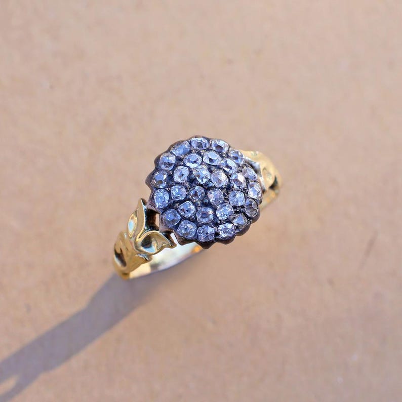 May include: A gold ring featuring a cluster of small, round, clear gemstones. The ring's band has a decorative floral design. The gemstones are set in a dark metal, creating a contrast with the gold band. The ring is set against a neutral background.
